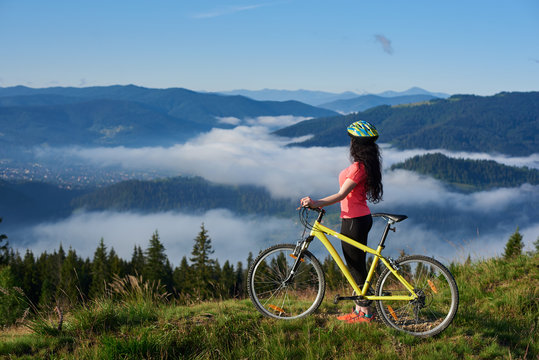 Side view of sporty female biker with yellow bicycle in the mountains, wearing helmet and red red t-shirt in the morning. Foggy mountains, forests on the blurred background. Outdoor sport activity