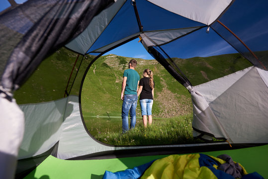 Back view of young pair resting near camping in the mountains at lake at sunny day. View from inside a tent