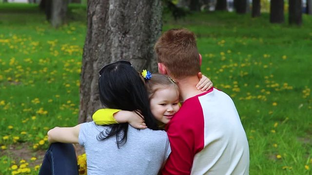 Cute funny little 4 year old daughter hugs and kisses her mom and dad outdoor. Happy family enjoys spring nature in city green park.