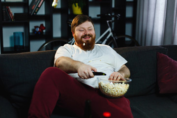 Fototapeta na wymiar Happy fat man eats pop-corn lying on the sofa before a table with beer