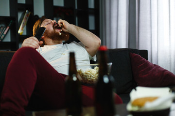 Happy fat man eats pop-corn lying on the sofa before a table with beer