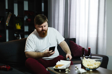 Fototapeta na wymiar Pretty fat man smiles checking his smartphone while he sits on the sofa and eats
