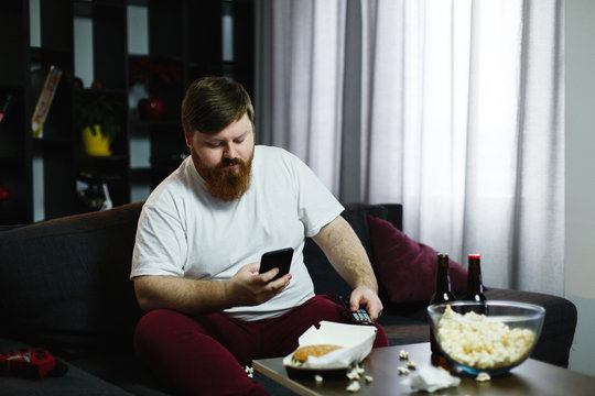 Pretty fat man smiles checking his smartphone while he sits on the sofa and eats