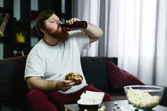 Fat man eats burger with beer sitting at the table before TV