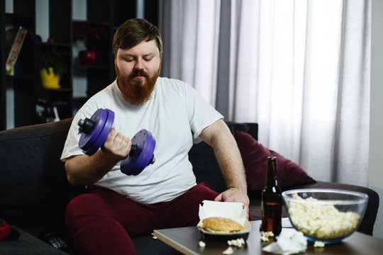 Fat man doing sport while he sits with food before a TV-set
