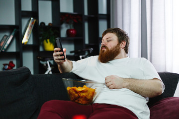 Pretty fat man smiles checking his smartphone while he sits on the sofa and eats