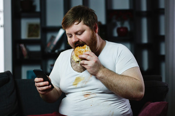 Ugly fat man checks something in his smarphone while he eats a burger
