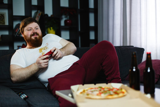 Happy fat man in dirty shirt plays video games drinking beer and eating pop-corn and chips