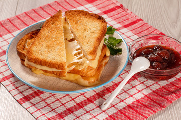 Toasted slices of bread with cheese and green parsley on white plate, bowl with strawberry jam and spoon on napkin