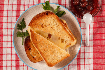Toasted slices of bread with cheese and green parsley on white plate, bowl with strawberry jam and spoon on napkin