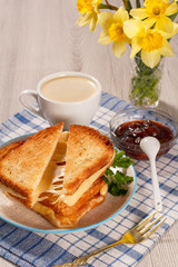 Toasted slices of bread with cheese and green parsley on white plate, fork, cup of coffee, bowl with strawberry jam, spoon and yellow daffodils on napkin
