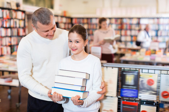 Portrait of tweenage girl with father standing in library with pile of books in hands