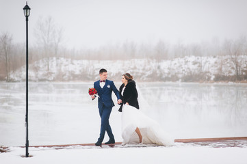 Young groom and bride walk on the snowy pier. The newlyweds are walking along the winter wharf, holding hands. Winter wedding.