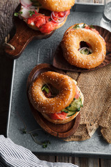 Bagels with salmon fish, cream cheese, cucumber and fresh radish slices on metallic tray on rustic gray wooden background