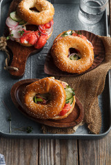 Bagels with salmon fish, cream cheese, cucumber and fresh radish slices on metallic tray on rustic gray wooden background