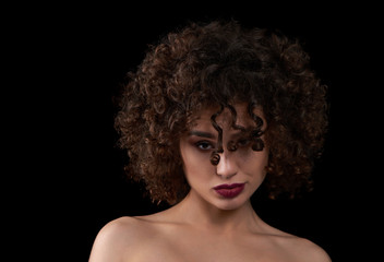 Exotic curly girl looking at camera and posing. Wearing light day make up with saturated violet lipstick. Having beatiful opened shoulders. Standing on black studio background.