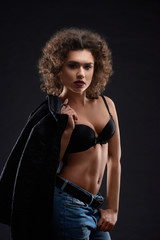 Fototapeta premium Sexy woman posing in black bra and blue jeans. keeping black jacket .Looking beatiful,confident, charismatic. Having pretty face features, many little curles and day make up. Studio black background.