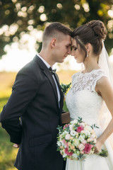 Stylish couple of happy newlyweds walking in field on their wedding day with bouquet. In the middle of the field ther is a big tree