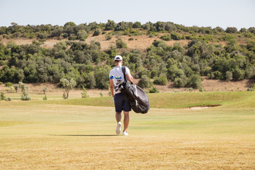 Golfer carrying his bag and clubs across a course.