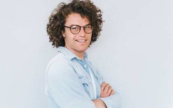 Horizontal profile view portrait of handsome smiling young male with curly hair, wears denim shirt and round trendy spectacles, looking to the camera. Copy space for advertisement. People race concept