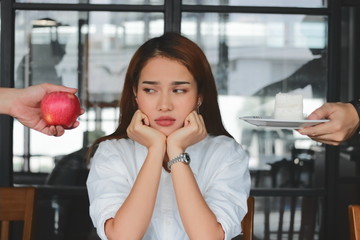 Pensive stressed Asian woman choosing between apple or unhealthy cake on the desk. Healthy lifestyle and dieting concept.  Selective focus and shallow depth of field.