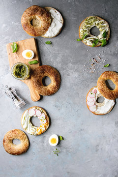 Variety of homemade bagels with sesame seeds, cream cheese, pesto sauce, eggs, radish, herbs served on crumpled paper with ingredients above over grey texture background. Top view, space.