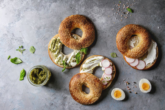 Variety of homemade bagels with sesame seeds, cream cheese, pesto sauce, eggs, radish, herbs served on crumpled paper with ingredients above over grey texture background. Top view, space.