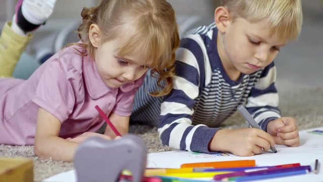 Medium shot of two little children drawing picture together using colored felt-tip pens and pencils when lying on the floor