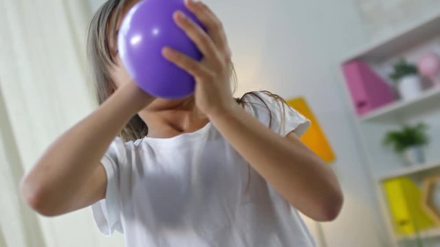 Medium shot of adorable little girl trying to blow up purple balloon, but then bursting out laughing