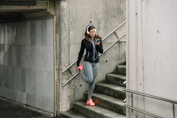 Sporty woman with headphones stretching legs for warming up before running in the city. Female athlete exercising and listening music with smart phone.