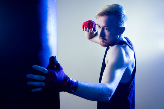 A young male boxer is doing sports in the gym. boxer, puts on boxing gloves on a dark background. the man strikes. red bandage on the hands