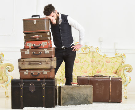 Baggage and relocation concept. Macho elegant on tired face, exhausted at end of packing, leans on pile of vintage suitcases. Man with beard and mustache packed luggage, white interior background.