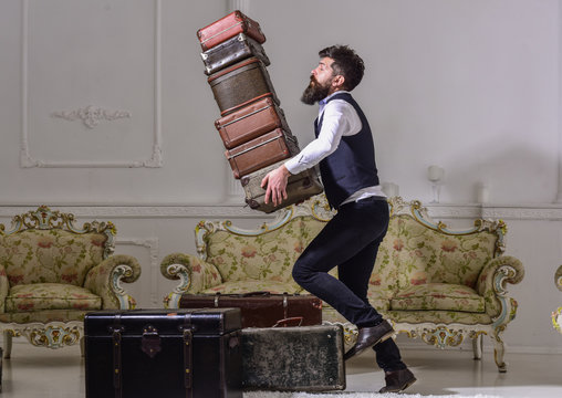 Macho elegant accidentally stumbled, dropping pile of vintage suitcases. Butler and service concept. Man with beard and mustache wearing classic suit delivers luggage, luxury white interior background