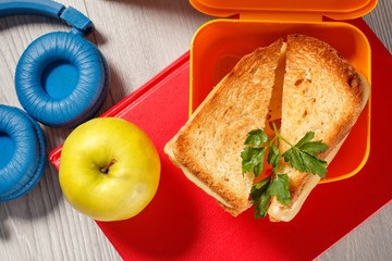 Yellow sandwich box with toasted slices of bread, cheese and green parsley, green apple, headphones and hardback books on the background