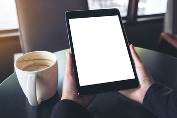 Mockup image of hands holding black tablet pc with blank white screen and coffee cup on table in cafe