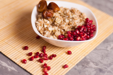 Cooked yummy warm oatmeal and heap of ripe fresh pomegranate seeds and three dried dates in white ceramic bowl on clean bamboo rug on worn gray scratched concrete with copy space