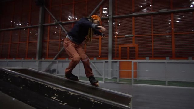 A man with long dreadlocks performs a grind on rollers in a skate park, slow motion
