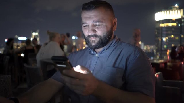 Young man texting on smartphone sitting in skybar at night
