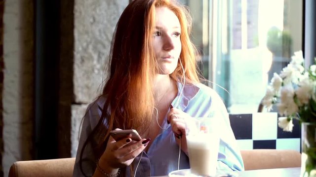 Pretty woman with red hair listening music on smartphone in cafe

