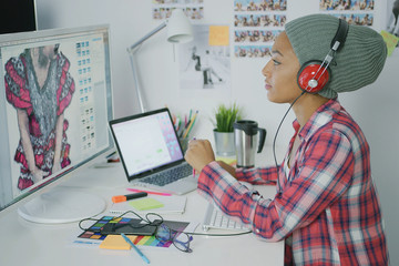 Hipster woman listening to music in headphones and designing clothing while working on computer at table in modern office.
