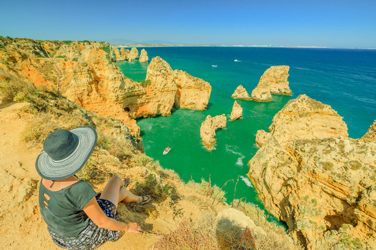Summer holidays in Algarve, Portugal. Lifestyle tourist with hat sitting on promontory of Ponta da Piedade and overlooks the coast of Lagos with iconic cliffs and limestone. Turquoise sea, sunny day.