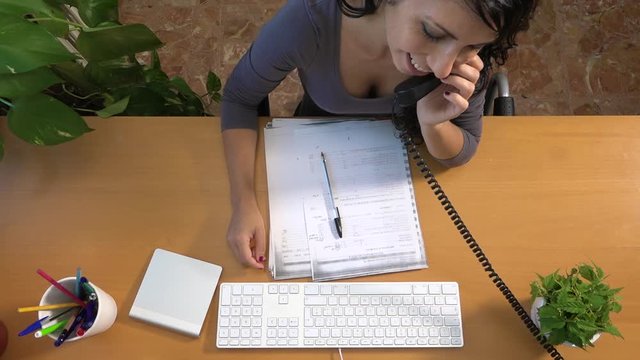 Caucasian woman talking on the telephone proofreading technical document draft. Businesswoman at office typing correction on the keyboard and picking up the landline. Brunette girl working on desk