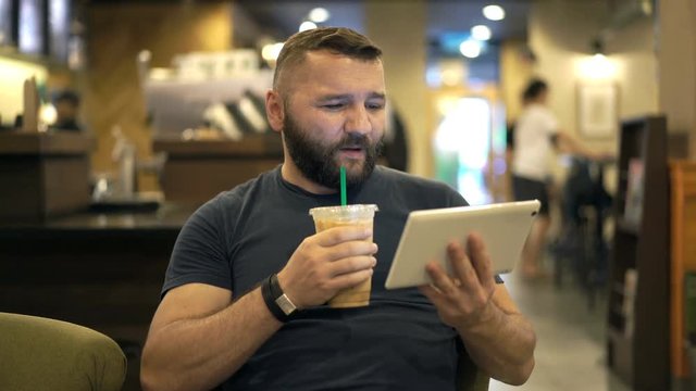 Happy man watching movie on tablet and drinking ice coffee in cafe
