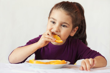 little cute child girl dressed in a purple blouse holding an orange and eating off a piece of orange