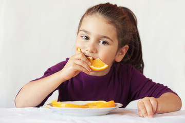 little cute child girl dressed in a purple blouse holding an orange and eating off a piece of orange