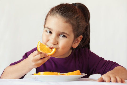 little cute child girl dressed in a purple blouse holding an orange and eating off a piece of orange