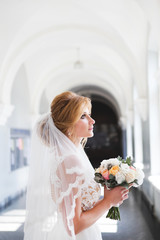 The bride in a beautiful wedding dress with a long veil stands in front of a large window and looks...