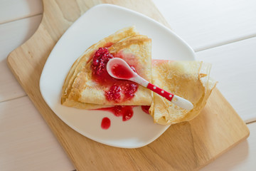 Pancakes with raspberry jam on a white plate stand on a natural wooden chopping Board and a light wooden table