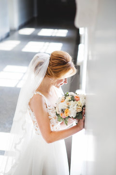  The bride in a beautiful wedding dress with a long veil stands in front of a large window and looks down. The bride's look down. Painting the shadow from the sun falls to the floor from the windows