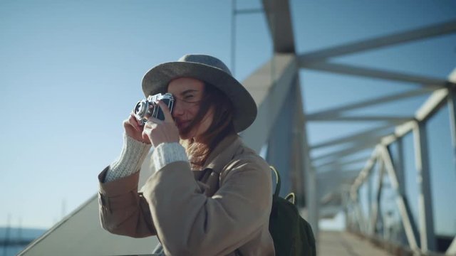 Cheerful hipster girl wearing trendy backpack and hat taking a photo of sea landscape on vintage film camera, young traveler woman walking in the city and capturing moments, Travel People Lifestyle